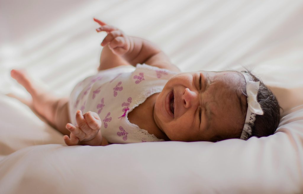 A close-up of a crying baby wearing a headband, lying on a soft bed indoors.